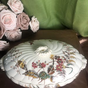 May include: A round, off-white ceramic tureen with a swirled, ribbed texture and a central floral knob. The lid features a colorful botanical print with red, yellow, and green flowers and ferns, complemented by decorative handles on each side.