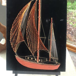 May include: A string art wall hanging featuring a sailboat design on a black velvet background. The ship has a solid terracotta-colored hull, two wooden masts, and sails created from intricate copper and gold-toned thread patterns.