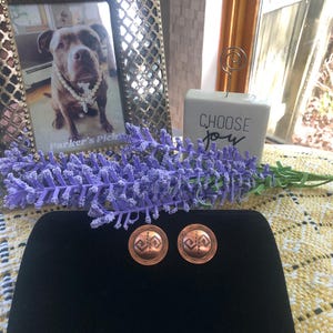 May include: A pair of round copper earrings featuring a central geometric pattern with a zigzag line and a scalloped border. The earrings have a warm metallic finish and are displayed on a black surface, showcasing a detailed engraved design.