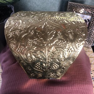 May include: A gold-colored, ornate box with a hinged lid. The box features an embossed floral and grape vine design across its surface. The box has a unique shape, wider at the top and tapering towards the bottom. The box is sitting on a maroon surface.