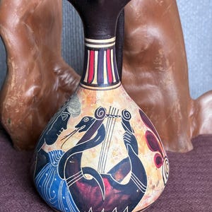 May include: A ceramic pitcher with a dark brown neck and a flared rim. The body features a painted scene of a figure in a blue garment playing a stringed instrument, rendered in black, red, and white against a mottled tan background.