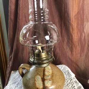 May include: A ceramic oil lamp with a speckled tan base, a side handle, and two textured vertical accents. The lamp features a brass-coloured metal burner assembly with a visible wick and a clear, bulbous glass chimney top for the flame.