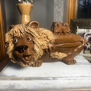 May include: A brown ceramic lion figurine with a detailed mane and facial features. Two small owl figurines sit on the lion's back. The lion is lying down, and the overall design is whimsical and decorative.