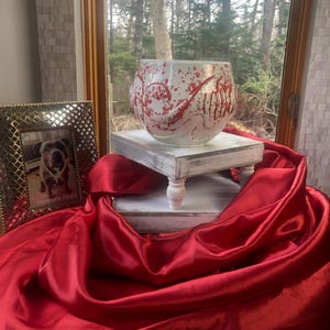May include: A white glass bowl with red splatter and swirl designs sits on a small, distressed white wooden pedestal. A red satin fabric drapes across the foreground. A framed photo of a dog is visible to the left.