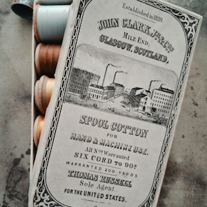 May include: A rectangular cardboard box of John Clark Jr. & Co. spool cotton featuring a vintage illustration of a factory. The box contains several wooden spools of thread in light blue, brown, and orange, alongside a small blue metal thimble.