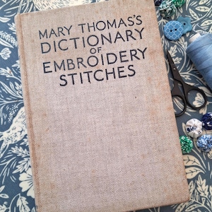 May include: A hardback book titled Mary Thomas's Dictionary of Embroidery Stitches with a beige fabric cover and black text. The book is positioned next to black metal scissors, a blue thread spool, and several small blue and green fabric yo-yos.