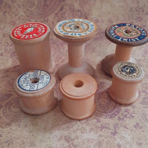 May include: A selection of antique wooden cotton spools with printed labels. The spools are in various sizes and styles, some cylindrical and others with a pedestal. The labels display text and graphics in red, blue, and gold.