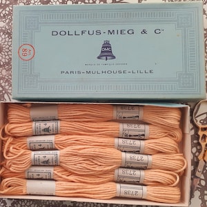 May include: A vintage box of peach-coloured embroidery floss with the text "DOLLFUS-MIEG & C". The box is light blue and has the text "PARIS-MULHOUSE-LILLE". Gold-coloured scissors are next to the box. The floss is labelled with the number "2738".