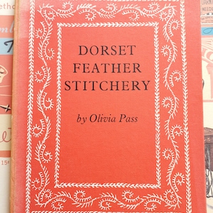 May include: A red book with a white decorative border and the title "Dorset Feather Stitchery" by Olivia Pass.