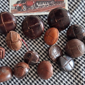 May include: A collection of round and square buttons in various shades of brown, featuring woven leather-like textures and smooth surfaces. Some buttons display a cross-stitched pattern on their faces, while others are plain and solid in appearance.