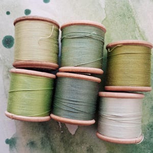 May include: Six wooden spools of thread arranged in a cluster, featuring various shades of green including olive, sage, moss, and a light greyish-green. Each spool has a smooth, cylindrical wooden core with visible thread wound around the centre.