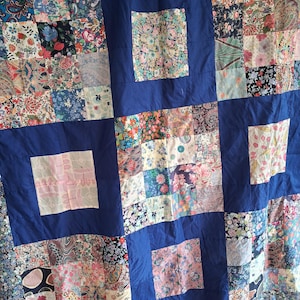 May include: A patchwork quilt featuring a grid of solid royal blue fabric squares interspersed with various floral and paisley patterned fabric squares in shades of pink, green, white, navy, and light blue, creating a multi-coloured textile design.