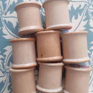 May include: A collection of eight cylindrical wooden spools arranged in a cluster. Each spool features a light tan, natural wood grain finish with a smooth surface, a central core, and raised circular rims at the top and bottom edges.