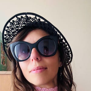 Pode incluir: Uma mulher usando uma faixa de cabelo de renda preta, óculos de sol pretos grandes, um colar gargantilha de crochê rosa e uma luva sem dedos combinando. Ela também está usando um suéter rosa claro. Os acessórios criam um visual elegante e feminino.