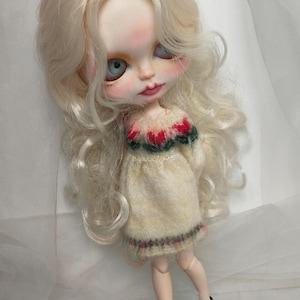May include: A doll with long, wavy blonde hair and blue eyes. The doll wears a cream-colored dress with a red and green floral pattern on the neckline and hem. Black Mary Jane shoes complete the look.