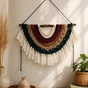 Puede incluir: Un tapiz de macramé semicircular colgado de una rama de madera oscura. Presenta capas concéntricas de cuerda anudada en color crema, beige, verde oscuro y burdeos, acentuadas con cuentas de madera natural y terminadas con borlas color crema.