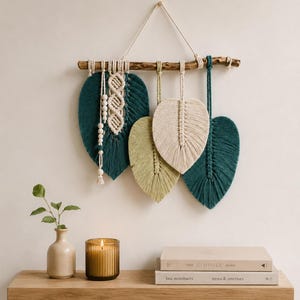 May include: A wall hanging featuring four macrame leaves in shades of dark teal, light green, and cream, suspended from a natural wooden branch. The leaves are crafted from cotton cord, with one leaf accented by a vertical row of wooden beads.