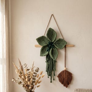 May include: A wall hanging featuring a large, five-petaled green macrame flower attached to a horizontal wooden dowel. Below the flower, green macrame fringe hangs down. A separate brown macrame leaf hangs from the same dowel by a cord.