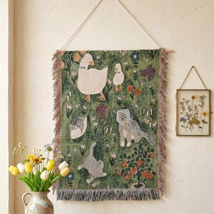 May include: A rectangular woven wall hanging featuring a green background with illustrations of white geese, a cat wearing a bonnet, and various colourful flowers and plants. The textile has fringed edges and is suspended by a light-coloured cord.