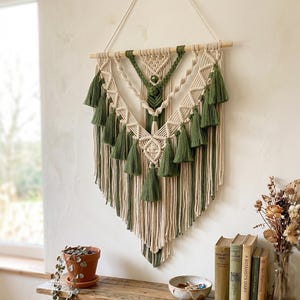 May include: A macrame wall hanging featuring cream and olive green cotton cord arranged in a V-shaped pattern. The piece is suspended from a horizontal wooden dowel and decorated with multiple olive green tassels along the edges.