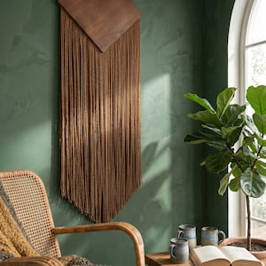 Puede incluir: Un adorno de pared marrón con una parte superior cuadrada de madera y largos flecos. La decoración de la pared se muestra contra una pared verde. Una silla tejida y una pequeña mesa de madera con tazas y un libro están en primer plano.