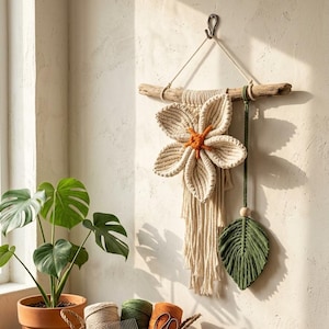 May include: A macrame wall hanging featuring a large cream-colored flower with an orange center, suspended from a piece of driftwood. A green macrame leaf hangs beside it. A basket contains yarn and a comb, with a sign reading Handmade Art - Sun Flower Macrame.