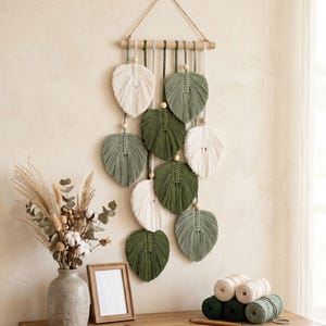 May include: A macramé wall hanging with leaf-shaped designs in white, green, and olive green. The leaves are suspended from a wooden dowel with wooden beads. The wall decor is displayed with a vase of dried flowers, a picture frame, and spools of yarn.