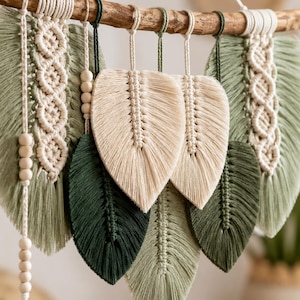May include: A macrame wall hanging featuring multiple leaf-shaped fibre elements in shades of sage green, dark forest green, and cream. The leaves are suspended from a natural wooden branch with cream-coloured cord and wooden beads.