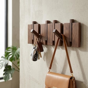 Op de afbeelding: Een houten wandkapstok met vijf haken. De kapstok is gemaakt van donkerbruin hout en heeft een modern design. Een sleutelbos hangt aan één haak en een bruine handtas aan een andere. De kapstok is aan een lichtgekleurde muur bevestigd.