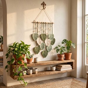 May include: A macrame leaf wall hanging in shades of green and cream hangs above a rustic wooden shelf. The shelf displays potted plants, mugs, and books. The room has a natural, bohemian aesthetic with neutral tones and textures.