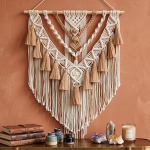 May include: A macrame wall hanging featuring cream and tan cotton cord, intricate knotted patterns, and multiple hanging tassels suspended from a wooden dowel. The design includes twisted rope elements and a central geometric knot structure.
