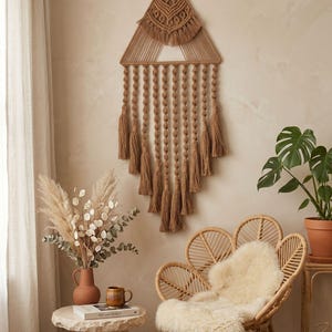 Puede incluir: Un tapiz de pared de macramé marrón con una parte superior triangular y borlas en cascada. La decoración de estilo bohemio se exhibe en una pared de tonos neutros. También se ve una silla tejida con una piel de oveja y una planta en maceta.