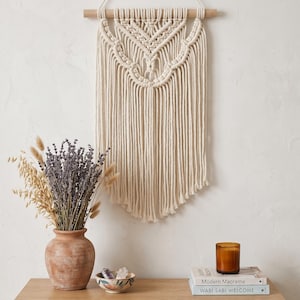 May include: A cream-colored macrame wall hanging with a wooden dowel, hanging against a white wall. A wooden cabinet with a woven front holds a terracotta vase with dried lavender and a small bowl with crystals. Books and a candle are on the cabinet.