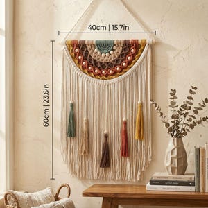 Puede incluir: Un tapiz de pared de macramé color crema, de 60 cm de largo y 40 cm de ancho, con un diseño de semicírculo en tonos marrones, amarillos y verdes. Cuenta con borlas colgantes de varios colores y cuentas de madera.