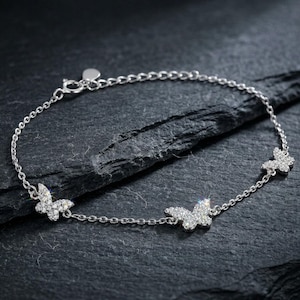 May include: A silver bracelet featuring three butterfly charms. Each butterfly is encrusted with small, sparkling stones. The bracelet has a delicate chain and a small heart-shaped charm near the clasp. The jewelry is displayed on a dark, textured surface.
