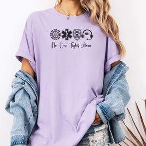 May include: A light purple t-shirt with black graphics of a fire department emblem, medical star, police badge, and 911 headset. The text "No One Fights Alone" is below the graphics. The shirt is worn with a denim jacket.