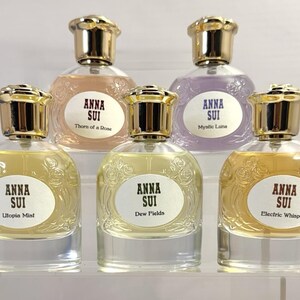May include: Six Anna Sui perfume bottles with gold-colored caps and labels. The bottles contain various colored liquids, including pink, purple, and yellow. The labels read "Thorn of a Rose", "Mystic Luna", "Utopia Mist", "Dew Fields", and "Electric Whisper".
