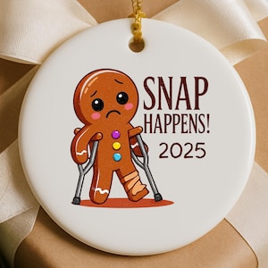 Könnte beinhalten: Weißer Keramik-Weihnachtsschmuck mit einem Lebkuchenmann mit gebrochenem Bein und Krücken. Der Text "SNAP HAPPENS! 2025" ist auf dem Ornament aufgedruckt. Mit goldener Aufhängeschnur.
