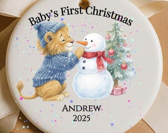 Puede incluir: Adorno navideño redondo con un cachorro de león con suéter y gorro azules, construyendo un muñeco de nieve. El adorno tiene el texto "Baby's First Christmas", el nombre "Andrew" y el año "2025". Cuelga de una cuerda dorada.