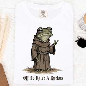 May include: White t-shirt featuring a cartoon frog in a brown robe with the text "Off To Raise A Ruckus". The frog has large eyes and is gesturing with one hand. The shirt has a Comfort Colors tag.