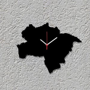 Könnte beinhalten: Eine schwarze Wanduhr in Form eines geografischen Umrisses, mit weißen Zeigern und einem roten Sekundenzeiger. Die Uhr ist an einer strukturierten, cremefarbenen Wand befestigt und erzeugt einen starken Kontrast.
