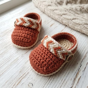 May include: A pair of handmade baby shoes. The shoes are crocheted in a rust-colored yarn with a cream-colored trim and a decorative strap. The strap features a chevron pattern in cream, rust, and orange. A wooden button secures the strap.