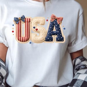 以下が含まれることがあります： 愛国的なデザインの「USA」という文字がプリントされた白いTシャツ。文字は赤、白、青の模様、リボン、星で装飾されており、祝日のお祝いやナショナルプライドを示すのに最適です。