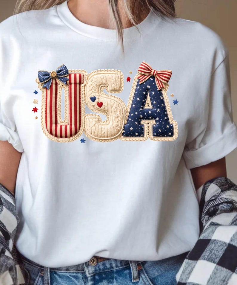 以下が含まれることがあります： 愛国的なデザインの「USA」という文字がプリントされた白いTシャツ。文字は赤、白、青の模様、リボン、星で装飾されており、祝日のお祝いやナショナルプライドを示すのに最適です。