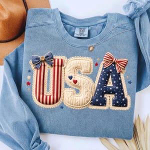 以下が含まれることがあります： 愛国的なデザインの「USA」という文字が入った水色のスウェットシャツ。文字は、リボンや星など、赤、白、青のアクセントで装飾されています。スウェットシャツは丸いネックラインと長袖です。