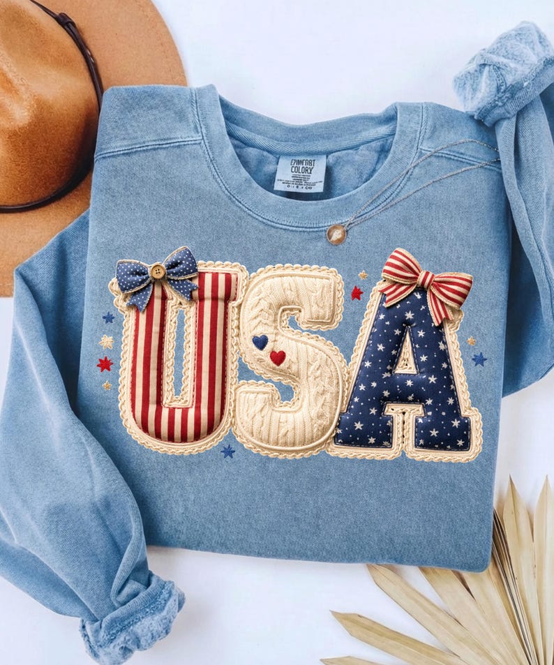 以下が含まれることがあります： 愛国的なデザインの「USA」という文字が入った水色のスウェットシャツ。文字は、リボンや星など、赤、白、青のアクセントで装飾されています。スウェットシャツは丸いネックラインと長袖です。