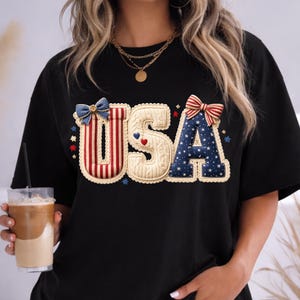 以下が含まれることがあります： 愛国的なデザインの「USA」という文字がプリントされた黒いTシャツ。「U」は赤と白のストライプ、「S」はクリーム色、「A」は白い星の付いた青色です。ゆったりとしたフィット感のTシャツです。