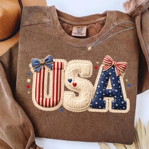 以下が含まれることがあります： 前面に「USA」の文字がアップリケされた茶色のスウェットシャツ。文字は、赤と白のストライプ、青い星、リボンなど、愛国的な模様で装飾されています。スウェットシャツはクルーネックと長袖です。
