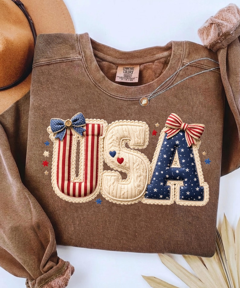 以下が含まれることがあります： 前面に「USA」の文字がアップリケされた茶色のスウェットシャツ。文字は、赤と白のストライプ、青い星、リボンなど、愛国的な模様で装飾されています。スウェットシャツはクルーネックと長袖です。