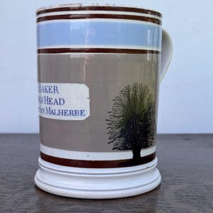 May include: A vintage ceramic mug with a handle, featuring a tree silhouette and text that reads "BAKER'S HEAD, TON MALHERBE." The mug has brown, blue, and white bands around the top and bottom, with a gray central section.